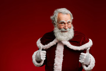 Waist up portrait of cheerful Santa Claus showing thumbs up while standing against red background,...