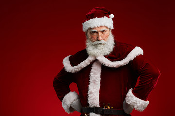 Waist up portrait of frowning Santa Claus standing with hands on hips while posing against red background in studio, copy space