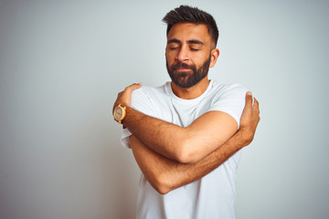 Young indian man wearing t-shirt standing over isolated white background Hugging oneself happy and...