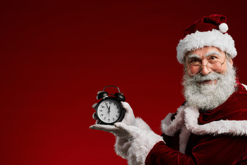 Waist up portrait of smiling Santa Claus holding clock while posing against red background in...