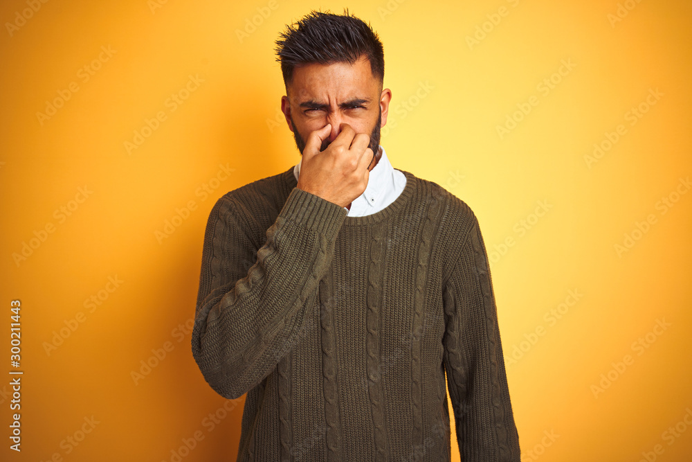 Canvas Prints Young indian man wearing green sweater and shirt standing over isolated yellow background smelling something stinky and disgusting, intolerable smell, holding breath with fingers on nose. Bad smell