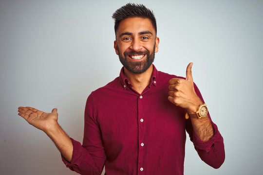 Young indian man wearing red elegant shirt standing over isolated grey background Showing palm hand and doing ok gesture with thumbs up, smiling happy and cheerful