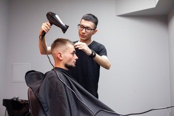 handsome guy has a haircut in a barbershop, a Kazakh barber dries a client's hair dryer against a gray wall, a hairdresser makes a short haircut