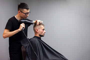 handsome guy has a haircut in a barbershop, a Kazakh barber dries a client's hair dryer against a gray wall, a hairdresser makes a short haircut