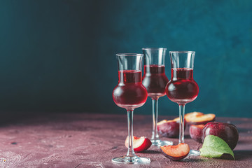 Plums strong alcoholic drink in grappas wineglass with dew. Hard liquor, slivovica, plum brandy or plum vodka with ripe plums on dark blue and  claret bordeaux concrete surface - obrazy, fototapety, plakaty