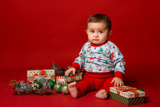 baby in Santa hat and Christmas pajamas sits with gift boxes on red background isolate