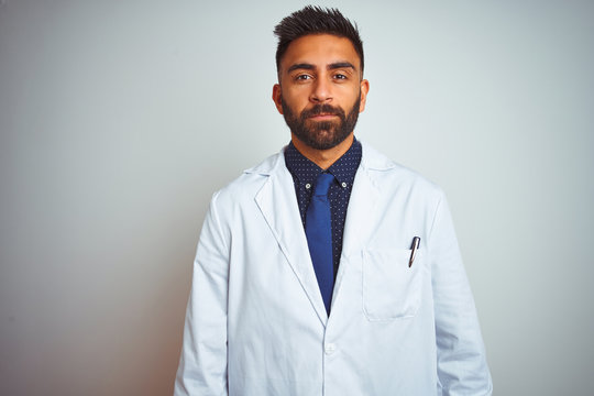 Young Indian Doctor Man Standing Over Isolated White Background With Serious Expression On Face. Simple And Natural Looking At The Camera.