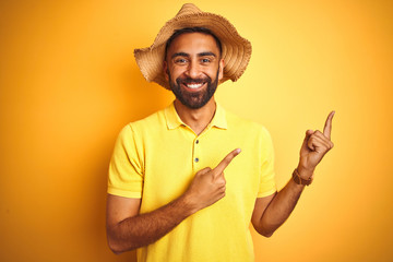 Young indian man on vacation wearing summer hat standing over isolated yellow background smiling...
