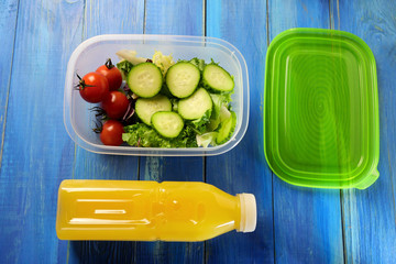 Plastic lunch box with fresh cucumber salad and cherry tomatoes on blue wooden table. Nearby is plastic bottle of orange juice and green cap. Healthy vegetarian food concept. Top view.