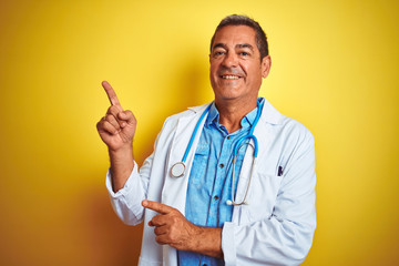 Handsome middle age doctor man wearing stethoscope over isolated yellow background smiling and looking at the camera pointing with two hands and fingers to the side.