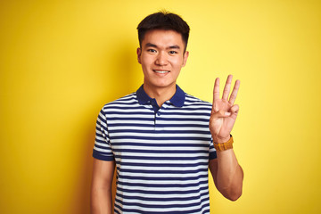 Fototapeta premium Young asian chinese man wearing striped polo standing over isolated yellow background showing and pointing up with fingers number three while smiling confident and happy.