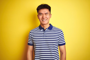 Fototapeta premium Young asian chinese man wearing striped polo standing over isolated yellow background winking looking at the camera with sexy expression, cheerful and happy face.