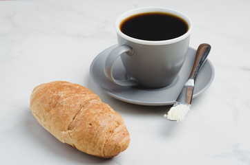 Coffee break with croissant, butter knife and cup of black coffee on a white stone table.