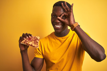 African american man eating slice of pizza standing over isolated yellow background with happy face...