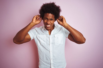 Young american man with afro hair wearing white shirt standing over isolated pink background covering ears with fingers with annoyed expression for the noise of loud music. Deaf concept.