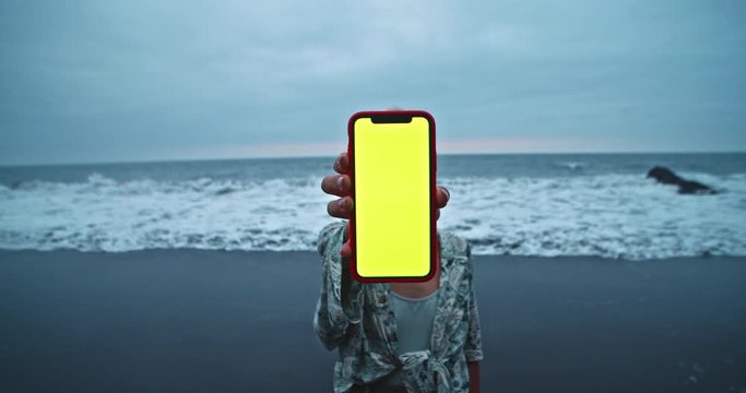 Woman showing vertical smartphone with blank chroma key greenscreen at sea. Female tourist holding mobile phone staying on the beach at calm blue ocean.