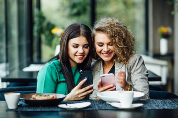 Fototapeta premium Young girls friends spending time together drinking coffee in the cafe, having breakfast and dessert. Youth, lifestyle, communication concept, using their phones.