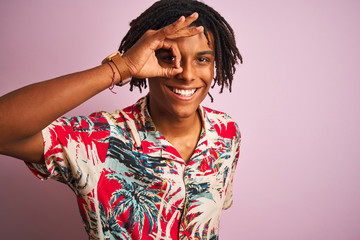 Afro man with dreadlocks on vacation wearing floral shirt over isolated pink background doing ok gesture with hand smiling, eye looking through fingers with happy face.