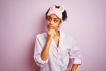 Young african american woman wearing pajama and mask over isolated pink background with hand on chin thinking about question, pensive expression. Smiling with thoughtful face. Doubt concept.