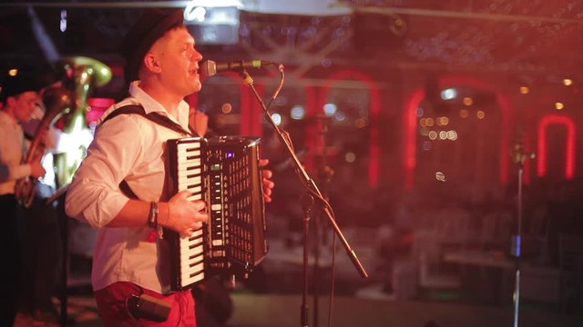 Performance of balkan folk jazz ensemble on stage. Man singing to microphone and playing accordion on blur colored background