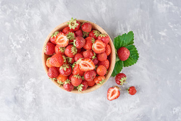 Fresh strawberries in a wooden bowl and green leaves on gray background, top view