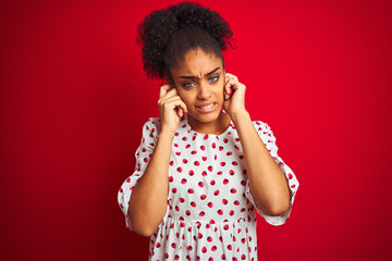 African american woman wearing fashion white dress standing over isolated red background covering ears with fingers with annoyed expression for the noise of loud music. Deaf concept.