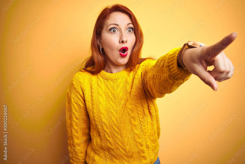 Wall mural Beautiful redhead woman wearing winter sweater standing over isolated yellow background Pointing with finger surprised ahead, open mouth amazed expression, something on the front