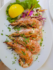 Grilled shrimp sprinkled with chopped herbs next to a slice of lemon and the green salad leaves on a white plate. Top view.
