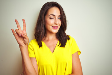 Young beautiful woman wearing yellow t-shirt standing over white isolated background smiling with happy face winking at the camera doing victory sign. Number two.