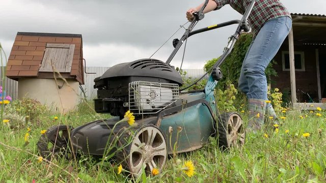 Blonde woman trying to start engine of a lawnmower, summer work in garden, cutting green grass