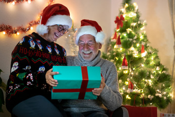 Christmas holidays at home. A senior couple smiling looking at one present still closed. Wearing Santa Claus hats. Love and serenity concept