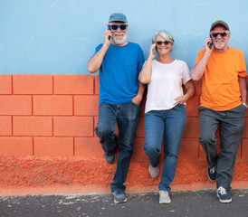 Three senior people with sunglasses and casual clothing using cellphone standing against an orange and blue wall. Complicity and friendship concept