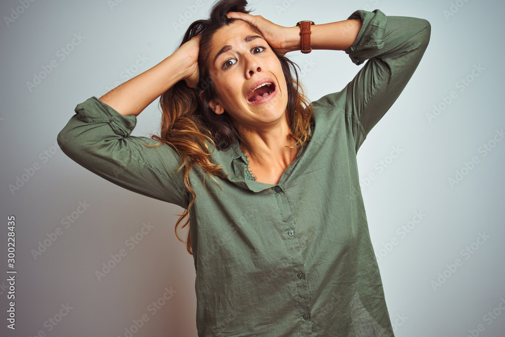 Wall mural Young beautiful woman wearing green shirt standing over grey isolated background Crazy and scared with hands on head, afraid and surprised of shock with open mouth