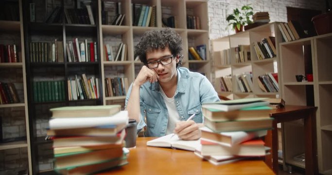 Asian student with curly hair is in library, sits at desk full of books. Guy wearing glasses preparing for exam and studying - education concept close up 4k