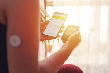 Woman comparing result of glucose level check on the traditional glucometer and on the mobile phone reading from modern technology remote sensor mounted on her forehand