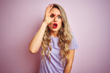 Young beautiful woman wearing purple t-shirt standing over pink isolated background doing ok gesture shocked with surprised face, eye looking through fingers. Unbelieving expression.