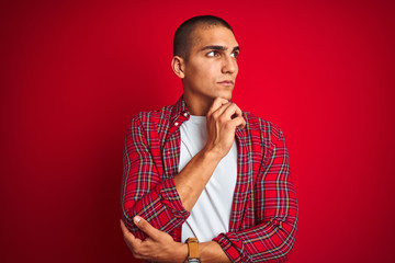 Young handsome man wearing a shirt using watch over red isolated background with hand on chin thinking about question, pensive expression. Smiling with thoughtful face. Doubt concept.