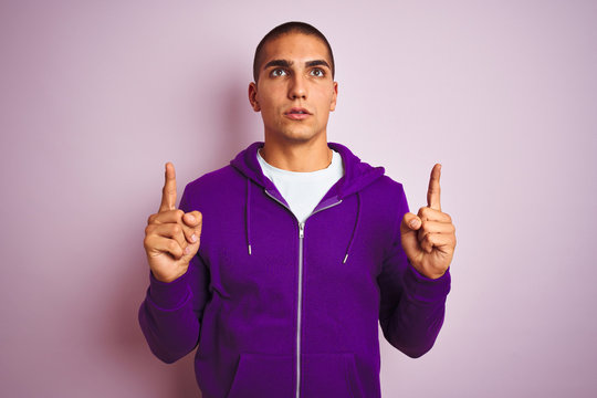 Young handsome man wearing purple sweatshirt over pink isolated background amazed and surprised looking up and pointing with fingers and raised arms.