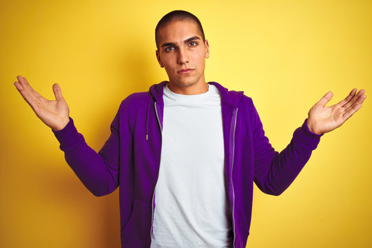 Young handsome man wearing purple sweatshirt over yellow isolated background clueless and confused expression with arms and hands raised. Doubt concept.