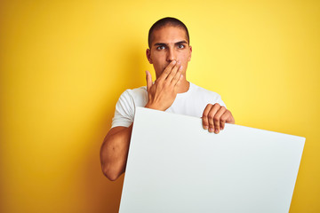 Young handsome man holding advertising banner over yellow isolated background cover mouth with hand shocked with shame for mistake, expression of fear, scared in silence, secret concept