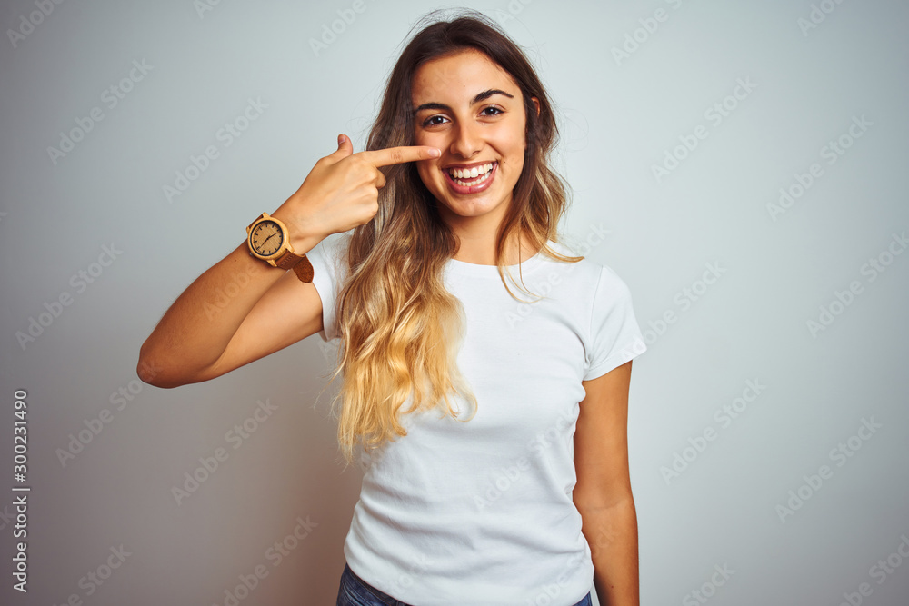 Poster Young beautiful woman wearing casual white t-shirt over isolated background Pointing with hand finger to face and nose, smiling cheerful. Beauty concept