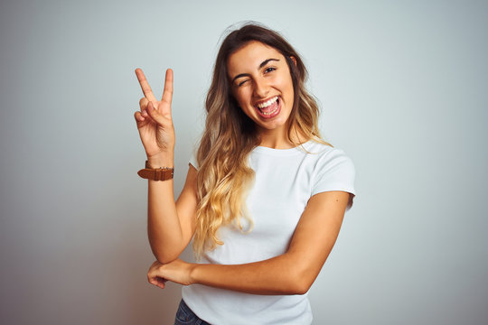 Young beautiful woman wearing casual white t-shirt over isolated background smiling with happy face winking at the camera doing victory sign with fingers. Number two.