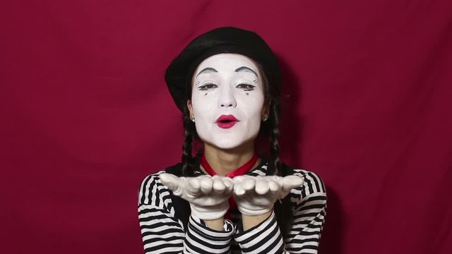 Beautiful girl mime sends air kiss and smiles looking at the camera. Beautiful girl in the image of a mime sends air kiss looking at the camera. Portrait on a red background.