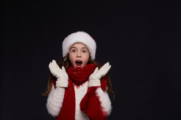 Portrait of young beautiful excited girl in red Santa hat on a dark background. Xmas fashion model with long curly hair. Winter holidays, Christmas, New Year concept.