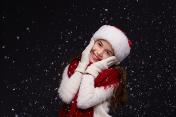 Portrait of young beautiful smiling girl in red Santa hat with white snowflakes on a dark background. Xmas fashion model with long curly hair. Winter holidays, Christmas, New Year concept
