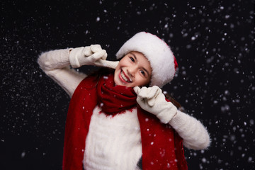 Portrait of young beautiful smiling girl in red Santa hat with white snowflakes on a dark background. Xmas fashion model with long curly hair. Winter holidays, Christmas, New Year concept