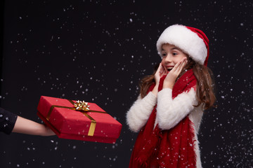 Happy exciting young beautiful smiling girl in white knitting pullover, red scarf and Santa hat get gift box on a dark background with snowflakes.Winter holidays, Christmas, New Year concept.