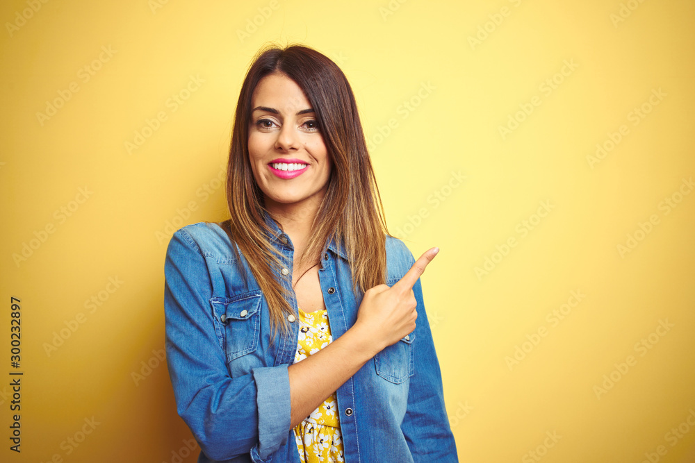 Poster Young beautiful woman standing over yellow isolated background cheerful with a smile on face pointing with hand and finger up to the side with happy and natural expression