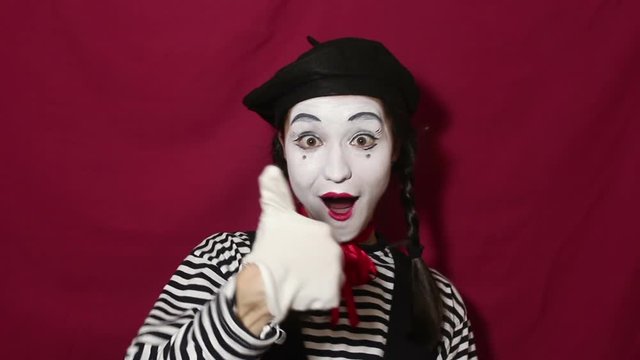 Beautiful girl mime smiles, shows thumb up and nods approvingly while looking at the camera. Beautiful girl in the image of mime shows thumb up while looking at the camera. Portrait on a red backgroun