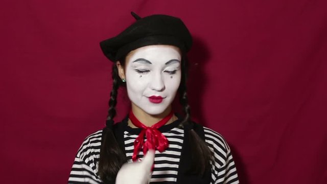 Beautiful girl mime smiles and winks eyes while looking at the camera. Beautiful girl in the image of mime winks and makes flirty movements with eyebrows looking at the camera. Portrait on a red backg